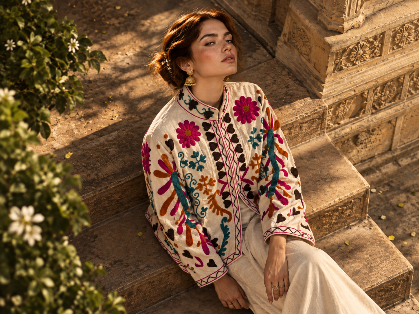 Seen from above, a model in the ivory Basant Panchhi jacket embroidered with pink blooms and dark hearts, seated on carved sandstone steps