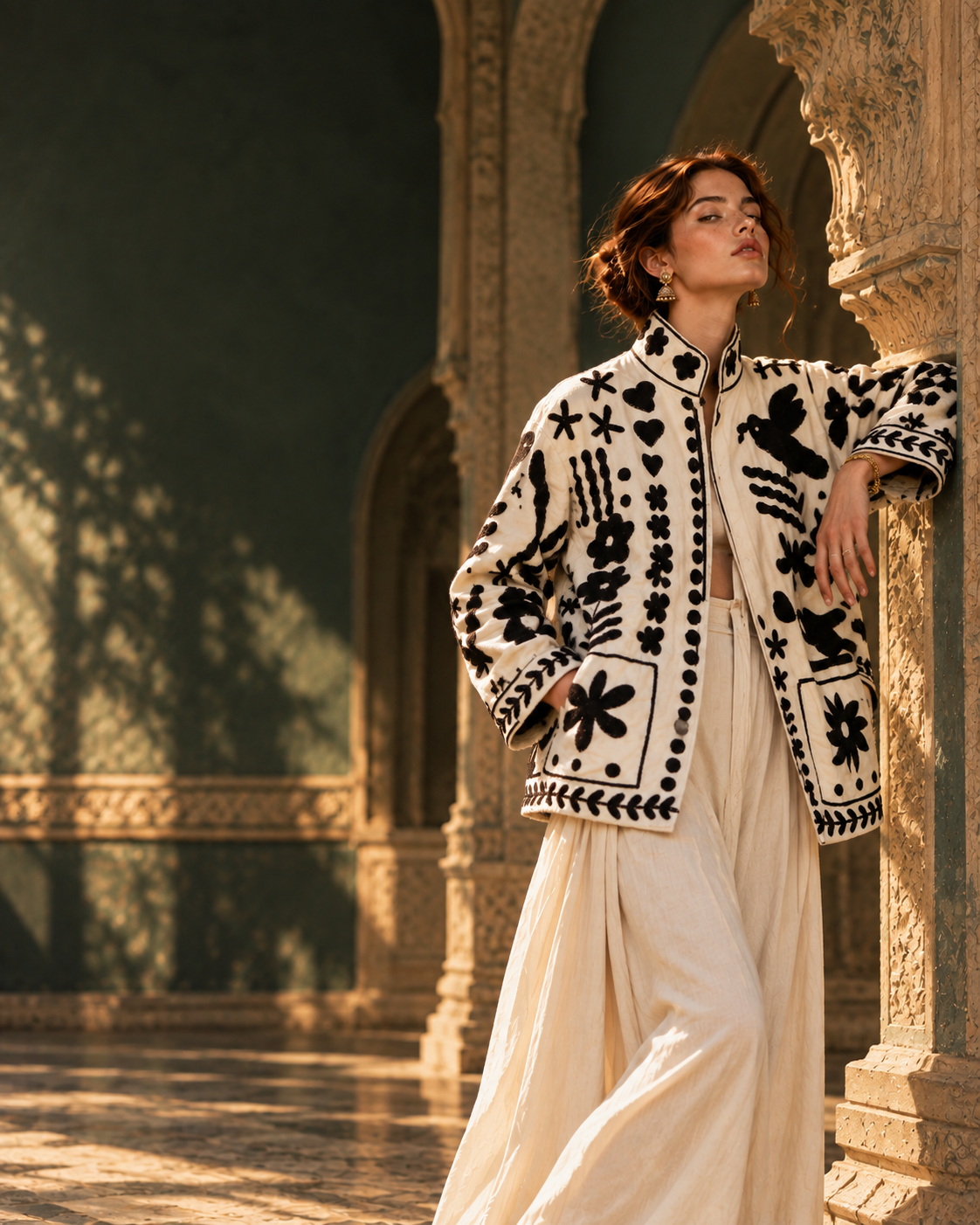 A model in the ivory Siyah Phool jacket with black motifs, leaning against a carved wall in a green-walled arcade